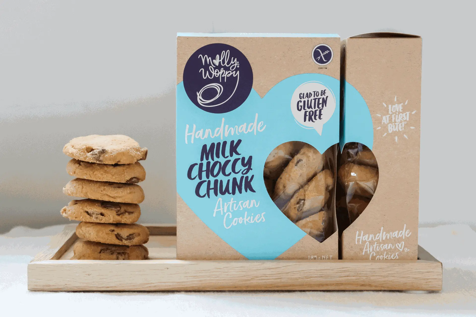 A box of Molly Woppy gluten-free Milk Choccy Chunk cookies next to a stack of cookies on a wooden tray, promoting handmade artisan quality.