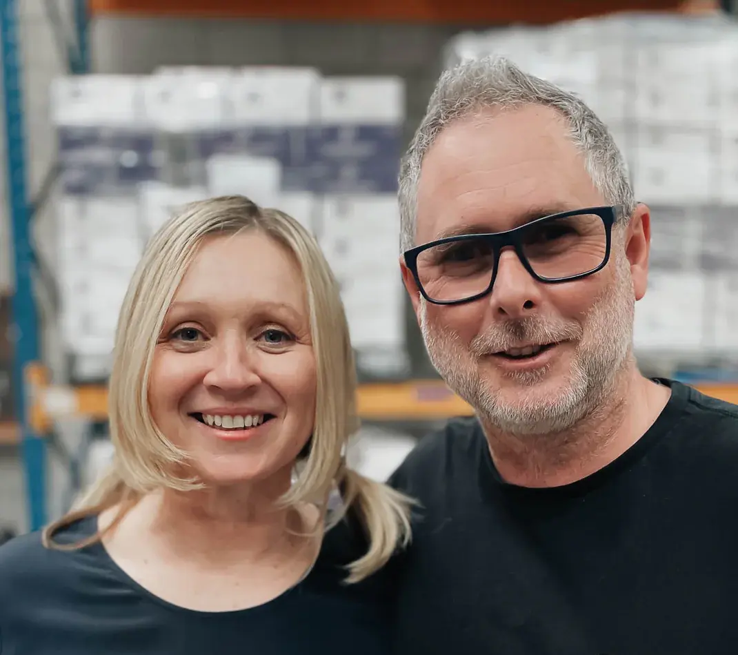 Smiling couple in Molly Woppy warehouse, with stacked boxes in background. Hayley Molloy with blonde hair in pigtails, Alistair Parker with gray hair, beard, and black glasses. Casual clothing, industrial storage environment.