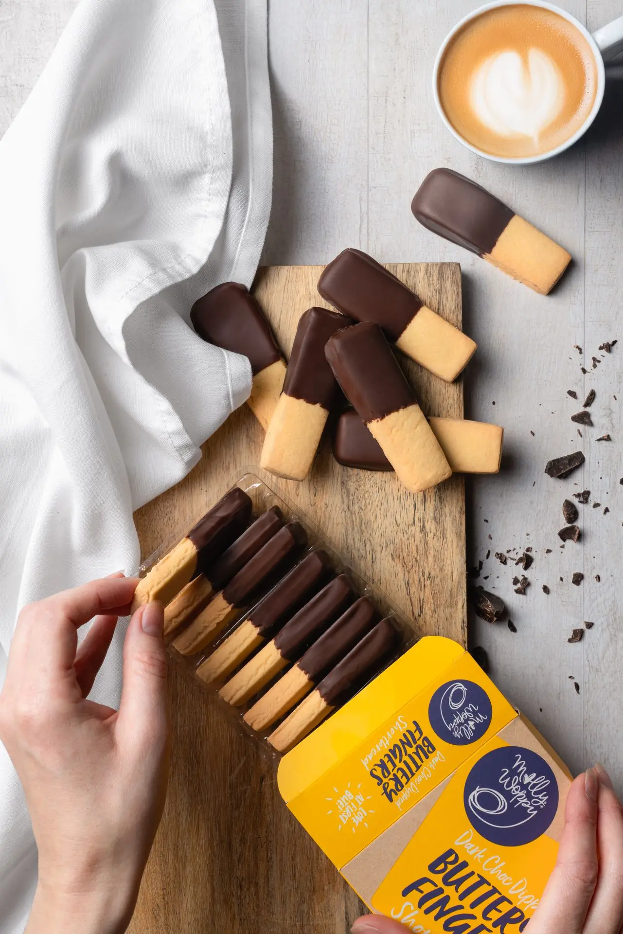 A hand reaches for a Molly Woppy chocolate-dipped butter finger cookie from yellow packaging, with scattered chocolate on a wooden board.