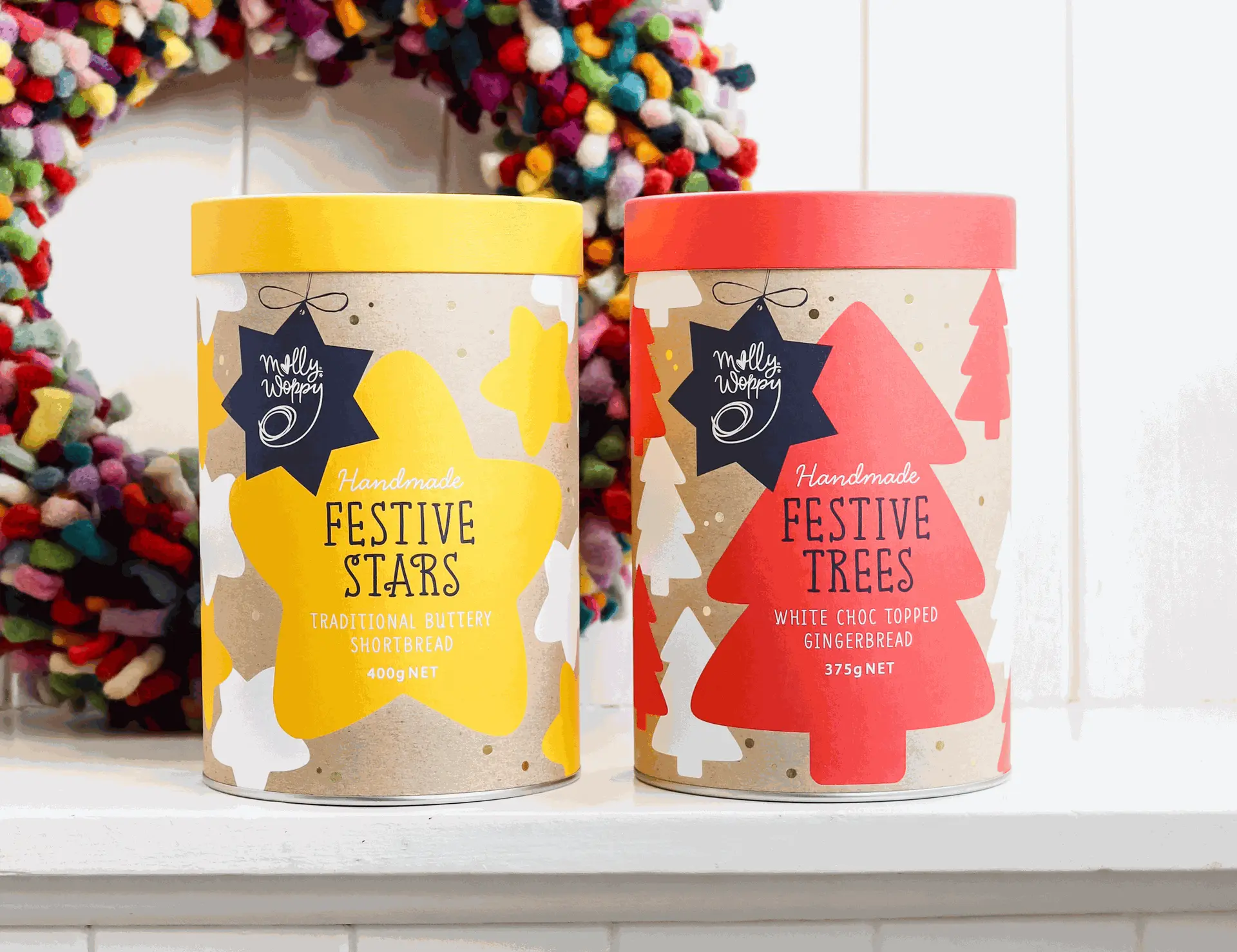 Two colourful festive treat containers: yellow Molly Woppy "Festive Stars" shortbread and red Molly Woppy "Festive Trees" gingerbread, against a vibrant wreath backdrop.