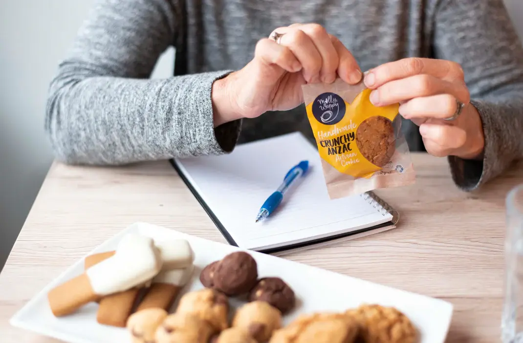 Business professional holding Molly Woppy Crunchy ANZAC cookie pack with plate of artisan cookies on desk — corporate cookie gifting and workplace treats in New Zealand.