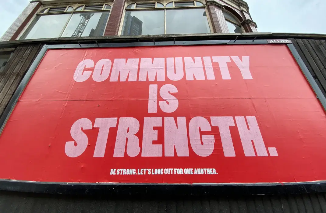 Red billboard with bold white text reading ‘Community is Strength’ — Molly Woppy highlights the importance of community and togetherness in New Zealand.