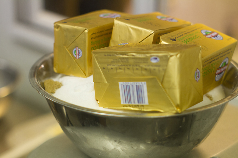 Metal mixing bowl filled with granulated sugar, light brown sugar, and several gold-wrapped blocks of unsalted butter, including labels reading “Unsalted Butter for Baking.” Ingredients are prepped for baking on a kitchen counter.