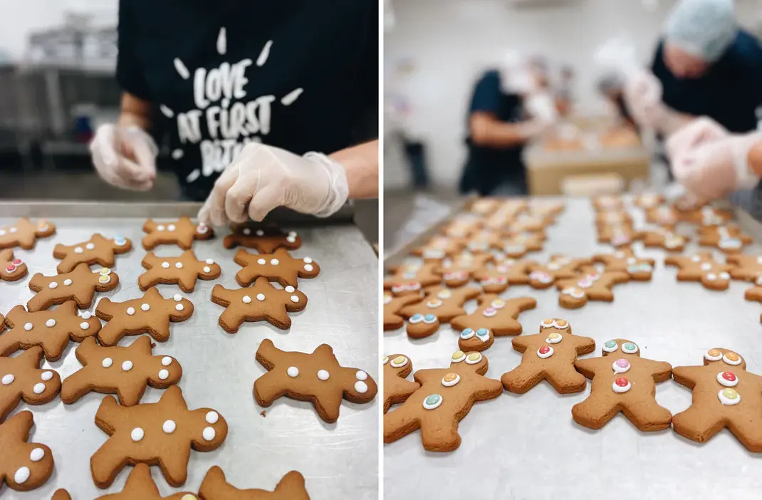 Molly Woppy Gingerbread cookies being decorated by gloved hands in a commercial kitchen. Workers in the background add candy eyes and buttons to gingerbread men on metal trays.